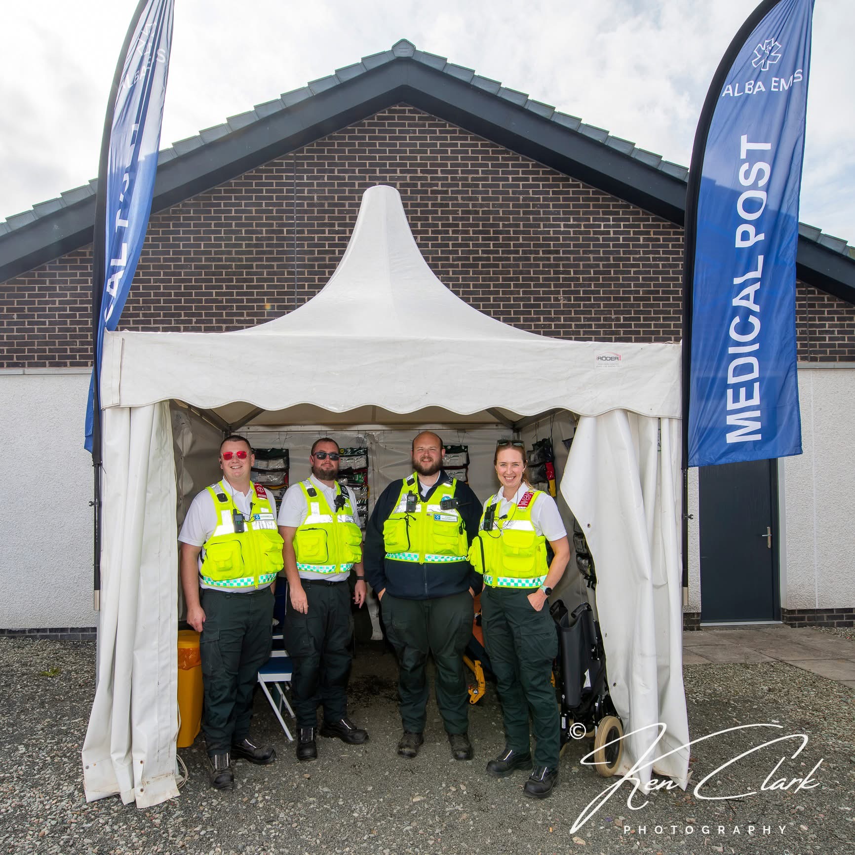 Four Alba EMS medics in high-visibility uniforms standing in the doorway of a white medical post tent with Alba EMS flags